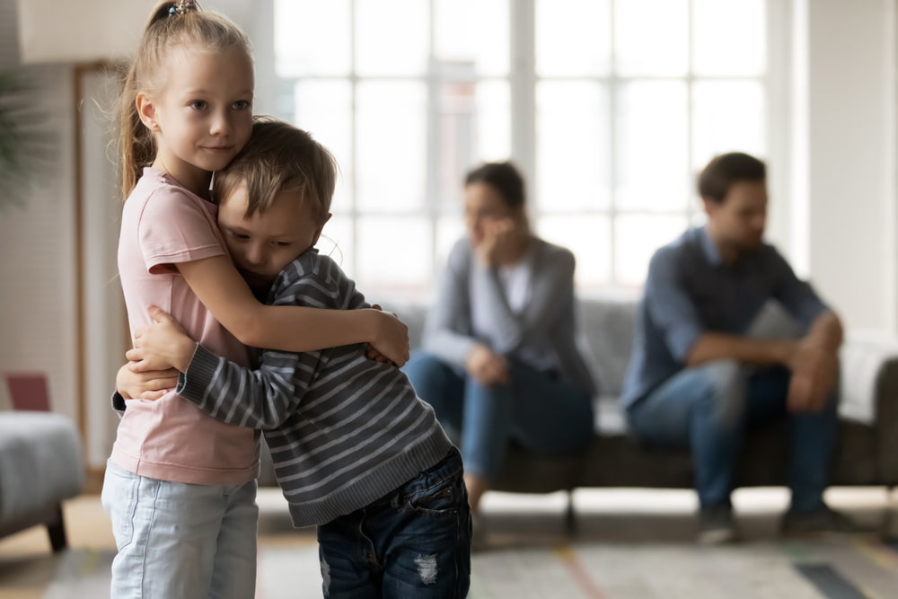Two young children hugging each other for comfort while adults sit in the background during a difficult family situation.